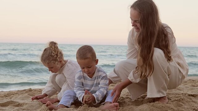 Mother and her two young children crouching and sitting on sandy beach playing with sand near water
