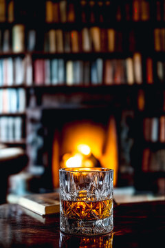 Glass of whiskey on a table in front of a cozy fireplace and bookshelves in a dim library room