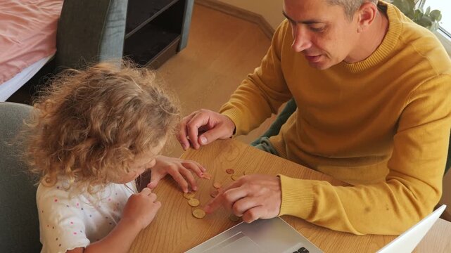 Father and little curly daughter counting euro coins on table together near laptop at home