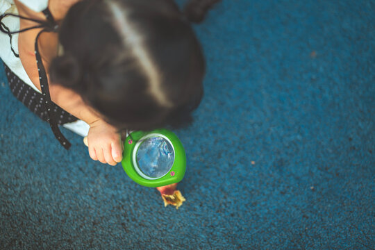 Curious toddler girl using magnifying glass to examine small natural object on ground outdoors, exploring environment through observation, discovery