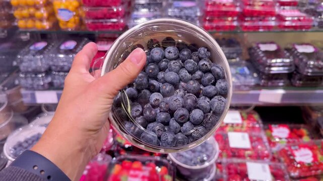 Hand Holding Packaged Berries in Supermarket &ndash; Raspberries, Blueberries, Strawberries and Cornelian Cherries Against Refrigerated Berry Display