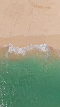 Aerial view directly above waves breaking on a sandy beach with emerald green ocean