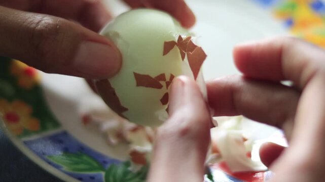 Close-up of hands carefully peeling off the eggshell membrane of a hard-boiled chicken egg on a decorative floral plate, a common morning ritual in Trinidadian and Caribbean kitchens 
