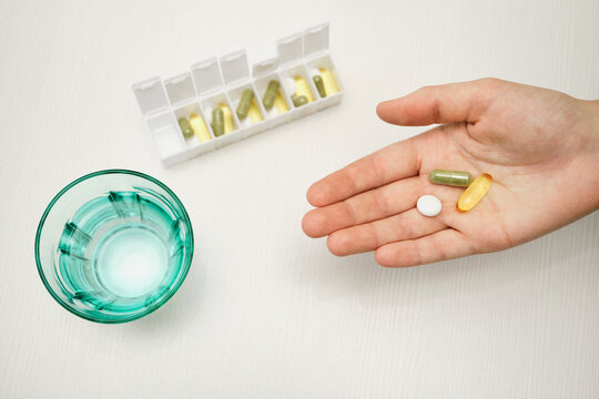 Hand holding medication with pill organizer and glass of water on white table