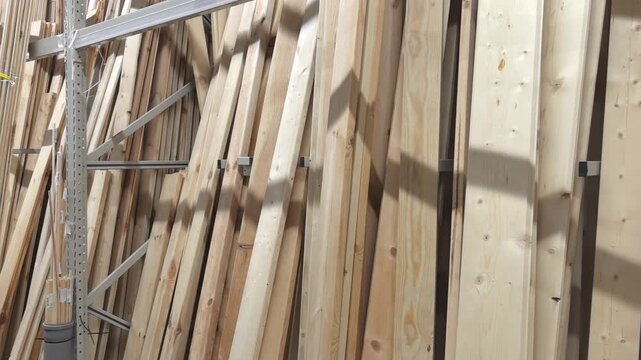 Wooden boards are neatly stacked in a spacious storage area. Light filters through, highlighting the textures of the wood. Shadows play across the surfaces, creating a dynamic visual.