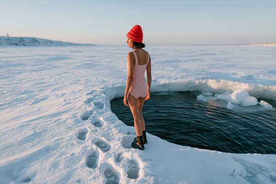 Woman in red beanie standing by a circular ice hole in a pale swimsuit at frozen shoreline for winter outdoor background and exploration poster