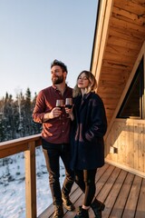 Fototapeta premium Couple on wooden cabin balcony holding wine glasses overlooking snowy forest for romantic retreat background and cozy lifestyle banner