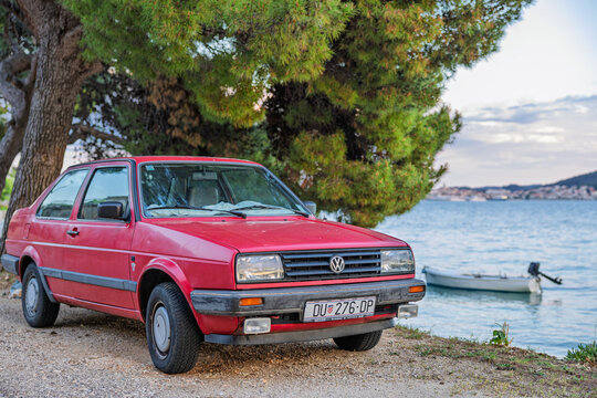 Red Volkswagen Jetta Vintage Car Parked By Seaside Under Pine Trees With Small Boat Nearby