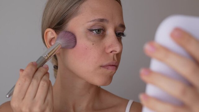 Young woman looks in a mirror while putting blush on her cheek with a makeup brush during daily makeup application
