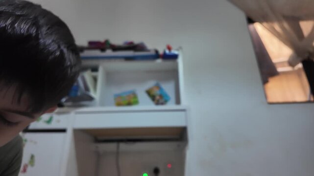 Child smiles at the camera while using a drill. He works on a white surface. Screwdriver bit is in the tool's head. The room holds shelves and toys. The child looks focused and cheerful