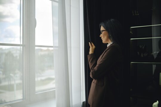 Woman in brown jacket gazes out window while reflecting on the day in a modern setting