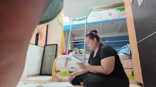 Woman reads documents while sitting on floor. Child's legs move nearby in child's room. Bedside storage and colorful bunk bed visible. Scene suggests home improvement planning. Footage captures quiet