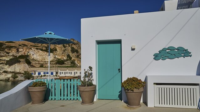 White building with turquoise door and view of a harbor in a Mediterranean landscape, Syrmata, boat garages, boathouse, boathouses, beach house, Goupa, Goypa, Goupa Kara, Kimolos, Cyclades, Greek Islands, Greece