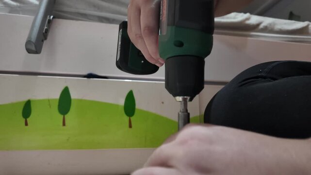Hand holds drill near child's room wall. Green trees decorate colorful wall background. Childhood room renovation in progress. Person works carefully to attach furniture
