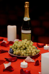 Obraz premium Green grapes on a plate with wine bottle and tealights on red tablecloth featuring scattered rose petals in romantic setting