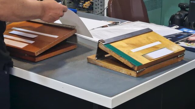 Close-up of hands aligning and trimming photographic sheets using an antique wooden guillotine paper cutter on a workshop table