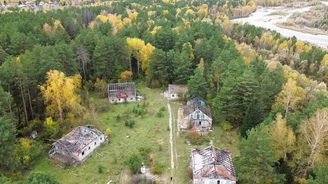 Aerial drone footage of an abandoned settlement surrounded by dense autumn forest, showing several deteriorating buildings and overgrown clearings, with a winding river and forested mountain slopes vi