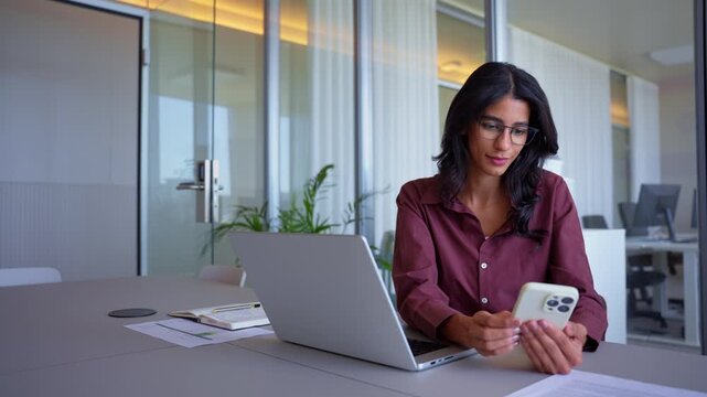 Laughing latin business woman in 30s reading funny message on mobile phone at office desk with laptop, young indian employee taking short break from busy work, medium shot of happy specialist.
