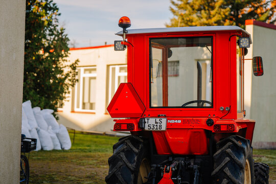 Red compact tractor with enclosed cab, triangular warning sign, roof beacon, large tires, and rear license plate parked beside a white building in an outdoor agricultural setting