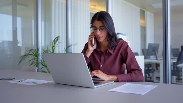 Busy indian business woman in 30s multitasking at office desk, talking on phone call, reading paper document and typing on laptop, latin manager doing paperwork, wide shot of employee at work.