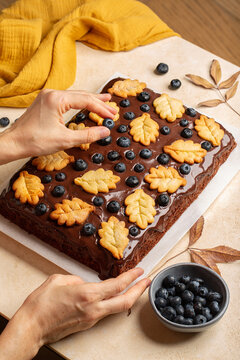 Homemade rectangular chocolate sheet cake covered with rich chocolate ganache and woman hands decorating the cake with yellow oak leaf cookies and fresh blueberries. Autumn-inspired cake