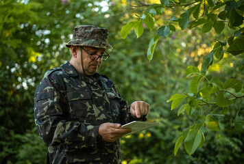 Camouflaged man uses tablet in woodland, tapping screen while studying its display. Uniformed field worker checks digital information beneath leafy branches with focused, serious expression. © Віталій Б.