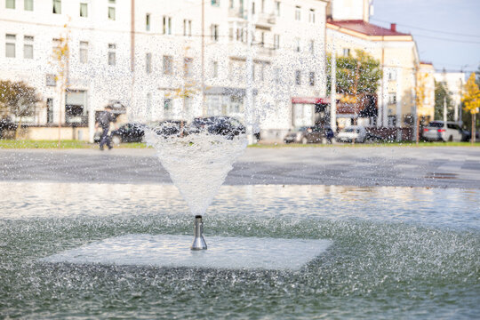 Close up of water fountain spraying jets into pool in urban square, with city buildings and traffic in blurred background