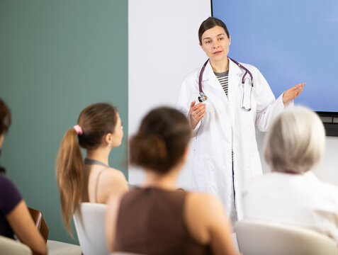 Positive young girl medical doctor dressed in white medical coat with stethoscope standing and talking in auditorium where sitting medical student different ages