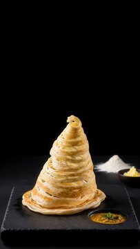 Fine dining presentation of Roti Canai flaky flatbread with dhal curry on a dark stone surface with dramatic negative space, featuring ghee and raw flour in the background.