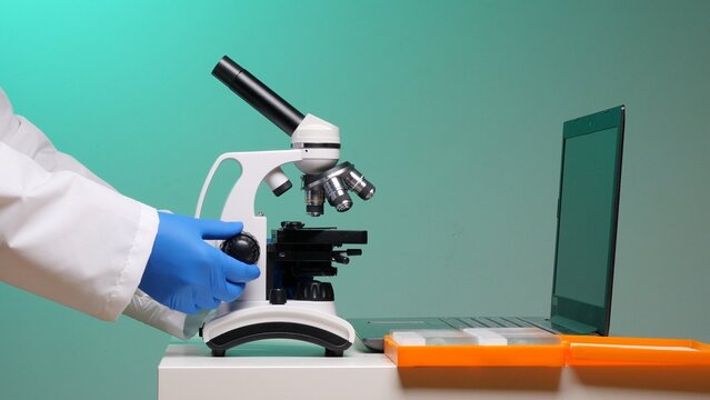 Scientist wearing a lab coat and blue gloves adjusting a microscope, performing research and analysis with laboratory equipment and a laptop in a scientific environment