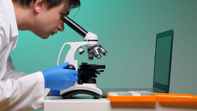 Scientist in white lab coat and blue gloves examining a slide under a professional microscope with laptop and slide box nearby, focused on medical and biological research and discovery