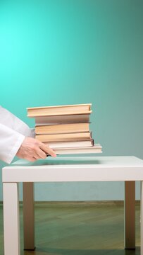 Hands wearing a white lab coat placing a stack of education books onto a clean white table, symbolizing learning, knowledge transfer, and academic study against a green background