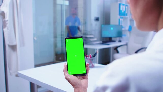 over-shoulder shot shows a doctor using a vertical smartphone with chroma key screen while a colleague exits behind, preserving mobile healthcare mockup value for screen replacement use.