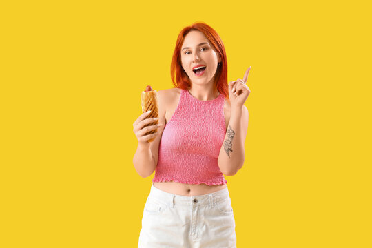 Fast food marketing and consumer choice, young woman holding a hot dog and pointing up with an excited expression on a bright yellow background