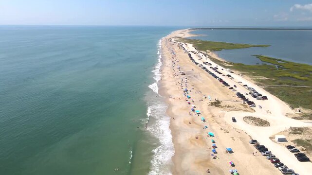 Rows of parked cars trace a narrow beach between ocean surf and marshy water.