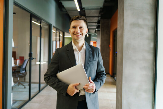 Professional man smiles confidently while holding a laptop in a modern office hallway during daytime