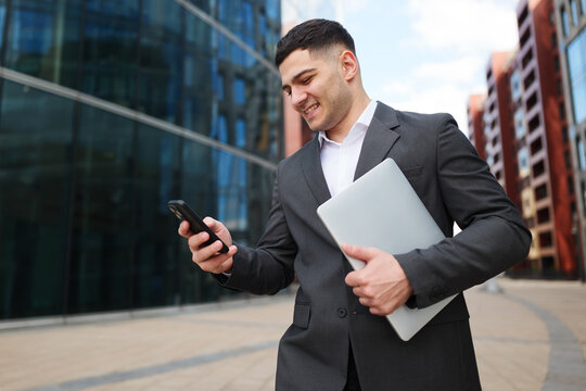 Business professional engaged in smartphone activity while holding a laptop near modern office buildings