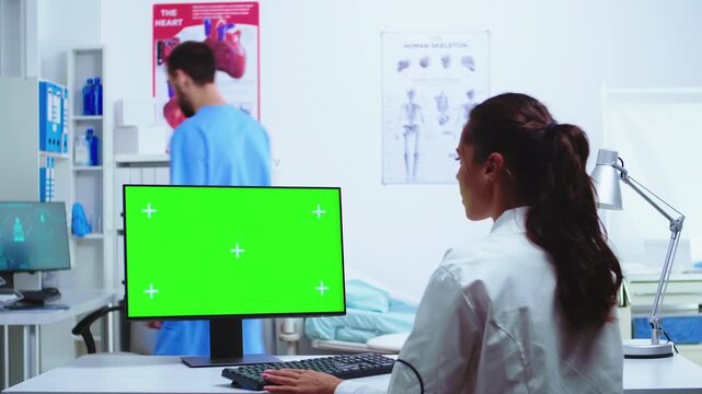 Doctor enters clinical notes at a green screen computer while an assistant in blue scrubs works nearby, showing healthcare documentation, medical records workflow and clinical technology use.