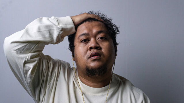 A compelling studio portrait showing an adult Asian male resting a hand on top of his head with a shocked and exhausted expression. The subject looks directly into the camera while