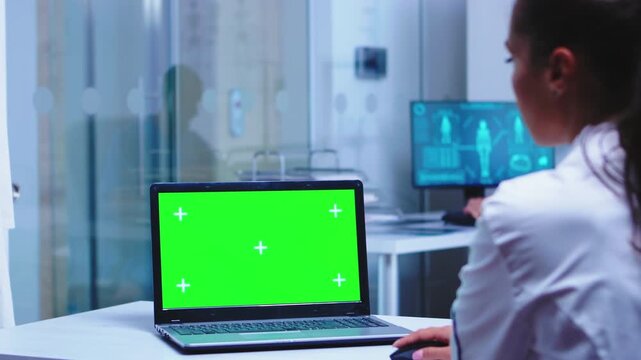 medical worker uses a chroma key laptop in the foreground while a colleague in scrubs opens the glass door, showing everyday clinic teamwork and digital workflow for healthcare mockup use.