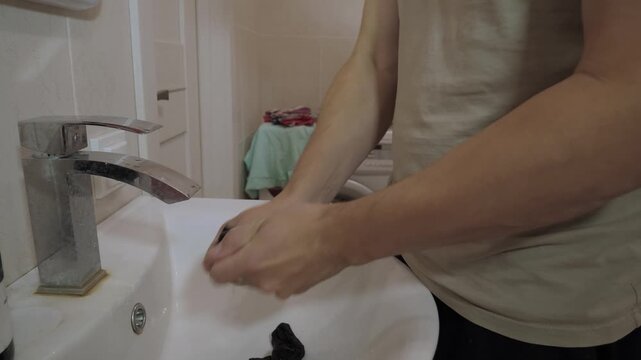 Male adult person squeezing dark sock by hand over white ceramic bathroom sink with chrome faucet