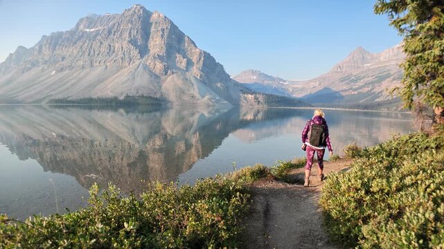 Hiker with backpack walks lakeside trail at Bow Lake, Banff National Park, as Rocky Mountains reflect on calm water beneath blue sky, serene wilderness journey in Banff National Park, Alberta, Canada.