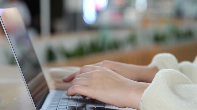 Close-up of hands typing on laptop keyboard. Person wears cozy white sweater. Background softly blurred, suggests indoor setting. Natural light illuminates scene. Focus remains on hands