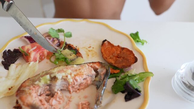 Hands cutting kiwi and salad with serrated knife beside partially eaten salmon and roasted sweet potato on white plate with yellow rim, low angled close-up at home dining table