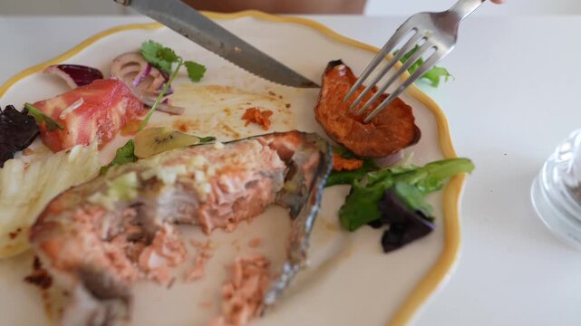 Close-up of fork and serrated knife cutting and lifting roasted sweet potato and salmon from scalloped white plate at dining table with tomato wedge and salad leaves