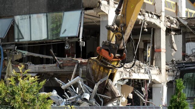 Demolition excavator with hydraulic crusher attachment tearing down concrete building facade, twisted metal ducts and rubble, yellow arm, glass windows, sunny day, no people, construction site