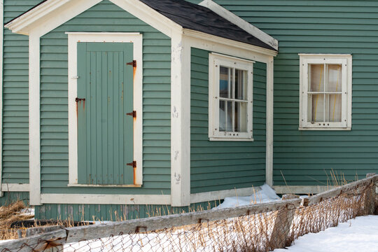 The porch section of a green wooden vintage building with white trim and asphalt shingles. There are two single-hung windows with curtains. The wooden door has three rusty metal hinges and a latch. 