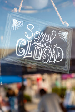 A small grey, square-shaped wooden business closed sign with white painted letters hangs in a glass window of a shop. The words sorry we're closed is handwritten in white chalk on the sign. 