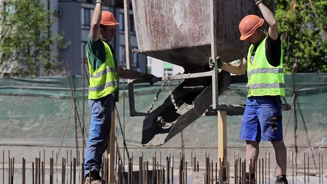 Two workers steady a concrete hopper above a steel rebar foundation, Laborers guide the crane bucket between exposed reinforcement rods outdoors