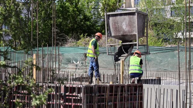 Construction workers position a crane bucket for concrete work, A suspended hopper is guided over formwork and rebar outdoors in direct daylight